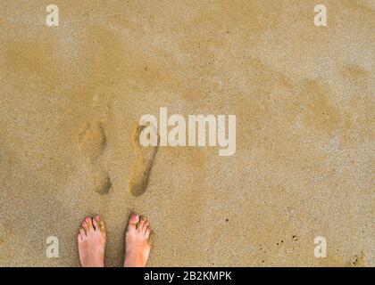 Pieds femelles barepieds avec une pédicure rouge à côté de leurs propres tirages sur le sable jaune humide. Vue de dessus. Banque D'Images