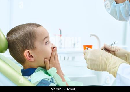 Portrait d'un enfant peur dans une chaise dentaire. Le garçon couvre sa bouche avec ses mains, craignant d'être examiné par un dentiste. Dentisterie pour enfants. Banque D'Images