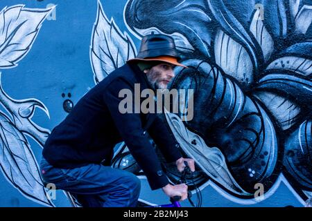 Un homme vêtu de cycles bleus passe devant un morceau d'art graffiti à motifs floraux bleus tout en portant deux chapeaux l'un sur l'autre. Banque D'Images