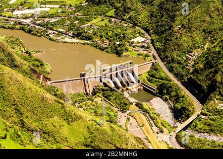 Une perspective aérienne montre un grand barrage hydroélectrique en béton enjambant une large rivière boueuse dans une vallée de montagne verdoyante. Le barrage comporte plusieurs vannes de déversoir et une structure de centrale électrique Banque D'Images
