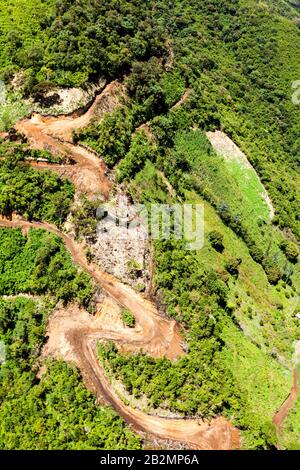 Une perspective aérienne montre un chemin de terre sinueux et non pavé coupant à travers une pente de montagne luxuriante et verdoyante. La route, apparaissant boueuse et érodée par endroits, serpente à travers une végétation dense, avec Banque D'Images