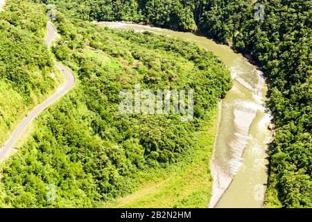 Une perspective aérienne montre une route courbe à deux voies longeant le bord d'une rivière large et peu profonde aux eaux vertes boueuses. La rivière coule à travers une vallée dense et verdoyante entourée de raides Banque D'Images