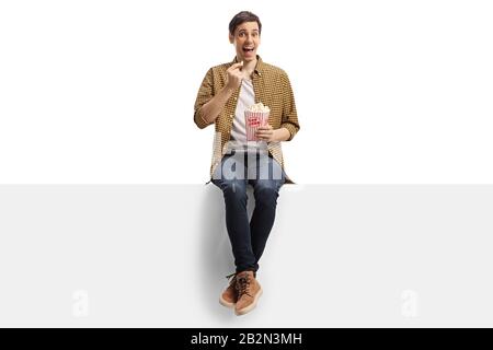 Un jeune homme heureux assis sur un panneau et manger du popcorn isolé sur fond blanc Banque D'Images