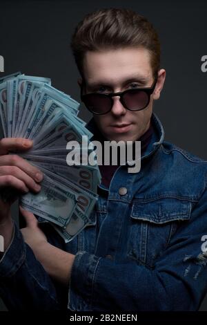 Portrait d'un jeune gars dans des lunettes de soleil et une veste denim regardant l'appareil photo tenant un ventilateur de billets de dollar. Sur fond gris. Revenu de l'argent conce Banque D'Images