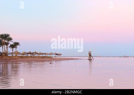 Plage vide au coucher du soleil rose en Egypte. Vue sur une belle plage de sable tropical vide avec parasols et lits de plage. Vue sur la mer calme Banque D'Images