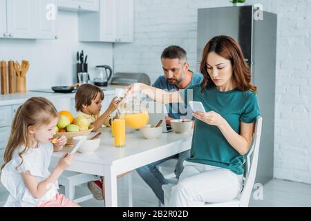 femme avec smartphone qui verse du jus d'orange dans le verre tout en étant assise près de la famille à l'aide de smartphones dans la cuisine Banque D'Images