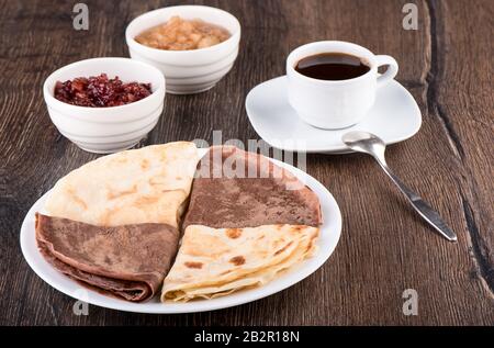 Les crêpes sont simples et au cacao. Crêpes sur une plaque blanche, confiture de pomme de différentes variétés et café sur un fond en bois. Russie Traditionnelle Banque D'Images