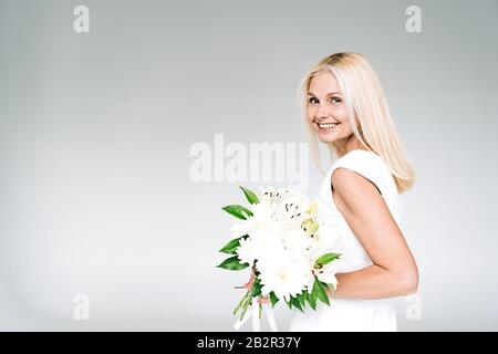 vue latérale d'une heureuse femme blonde mûre avec bouquet blanc isolé sur gris Banque D'Images