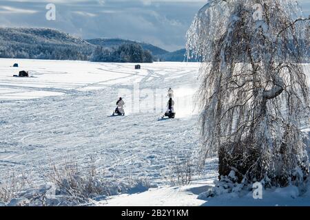 Trois motoneiges qui traversent la neige et la glace sur le lac Pleasant avec trois cabanes de pêche sur glace en arrière-plan près de Speculator, NY USA Banque D'Images