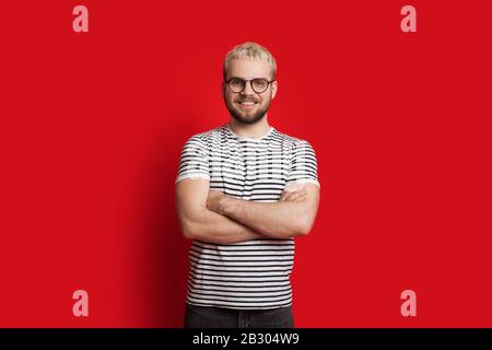 Homme caucasien confiant avec des cheveux blonds et une barbe courte se posant avec des mains croisées sur un fond rouge Banque D'Images
