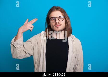 Un homme caucasien non rasé avec de longs cheveux gestuelle une photo dans sa tête tout en regardant à travers des lunettes sur un mur bleu Banque D'Images