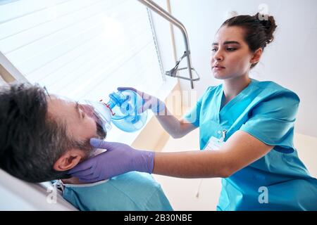 Vue en dessous de la jeune infirmière dans des gants chirurgicaux utilisant un masque de valve de sac tout en donnant de l'oxygène au patient à l'hôpital Banque D'Images