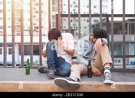 Deux jeunes hommes ivrent et s'assoient dormir dans la rue avec une bouteille de bière en mécontent. Banque D'Images