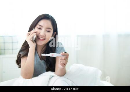 Belle jeune femme asiatique heureuse tenant le test positif enceinte dans la main et en utilisant un téléphone mobile intelligent avec le sourire tout en assis sur le lit à home.process Banque D'Images