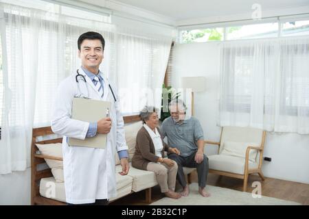 Jeune homme caucasien médecin debout main tenant dossier regarder l'appareil photo avec sourire et deux vieux vieux couple asiatique s'asseoir sur le canapé Banque D'Images
