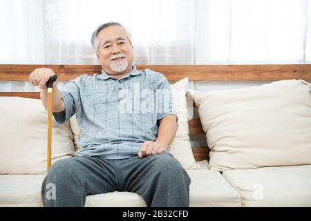 vieux homme âgé assis, reposant ses mains sur un bâton de marche en bois assis sur le canapé dans la salle de séjour de la maison après la retraite. Banque D'Images