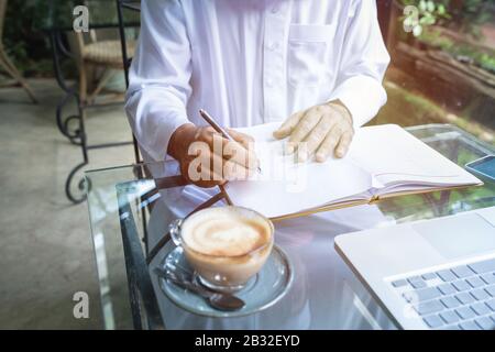 Jeunes hommes d'affaires arabes musulmans dans un café portant le hijab habillés écrire sur le fichier de livre avec ordinateur portable sur table.Focus sur la main. Banque D'Images