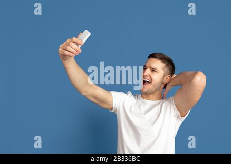 Prendre selfie. Portrait caucasien du jeune homme isolé sur fond bleu de studio. Magnifique modèle masculin dans un style décontracté, aux couleurs pastel. Concept d'émotions humaines, expression faciale, ventes, publicité. Banque D'Images