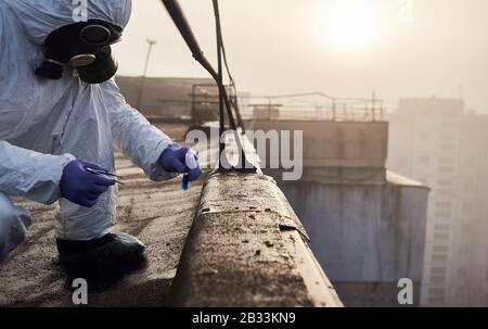 Cliché court d'un chercheur effectuant un test de laboratoire à l'aide de brucelles et de réactif bleu dans une fiole en verre et d'une pierre à l'extérieur, portant des vêtements de protection et un masque à gaz. Concept de pollution atmosphérique Banque D'Images