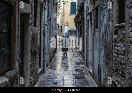 Vieille dame, courbée à l'aide d'un bâton de marche, marchant seul à travers les rues humides de Venise, Italie Banque D'Images