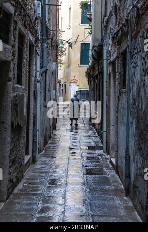 Vieille dame, courbée à l'aide d'un bâton de marche, marchant seul à travers les rues humides de Venise, Italie Banque D'Images