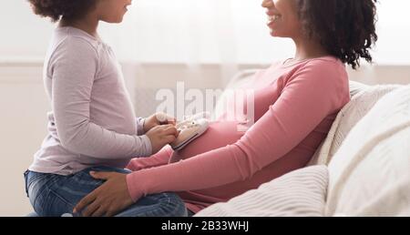 Petite Fille Mettant De Petites Chaussures Pour Bébé À La Ceinture De Mère Afro Enceinte Banque D'Images