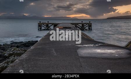 La vieille jetée de Portencross en Ecosse et les collines d'hiver sur l'île d'Arran tout comme le soleil descend produisant un spectaculaire coucher de soleil ciel. Banque D'Images