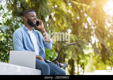 Joyeux Afro Guy Avec Ordinateur Portable Et Vélo Parler Sur Cellphone À L'Extérieur Banque D'Images