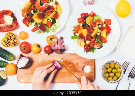 Processus de cuisson. Vue de dessus des mains féminines hachez l'oignon sur un tableau de découpe à côté des ingrédients et des assiettes pour salade de légumes. Banque D'Images