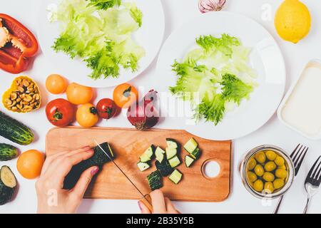 Cuisson d'aliments sains. Vue de dessus des mains de la femme couper le concombre frais à bord de la planche à découper à côté des ingrédients et des assiettes pour salade de légumes. Banque D'Images