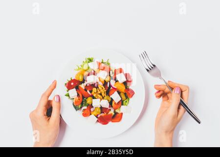 Les mains féminines tiennent la fourchette près de la plaque de salade grecque de légumes frais sur la table blanche, vue du dessus. Banque D'Images