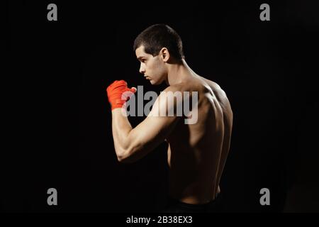 Boxeur sportif pratiquant un coup de pied latéral. Bandages rouges sur les mains. Boxeur musculaire sur fond noir. Concept sportif. Banque D'Images