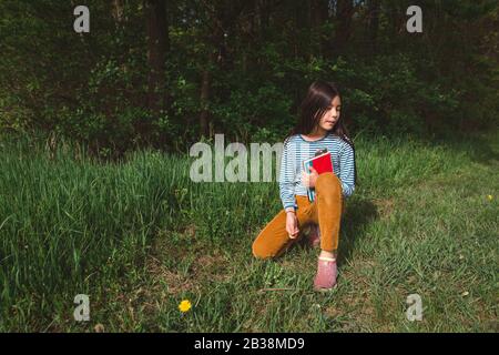 Une jeune fille paisible est assise dans une grande herbe en regardant avec un livre dans ses bras