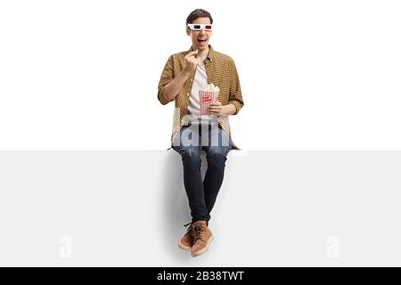 Jeune homme avec des lunettes en relief assis sur un panneau et manger du popcorn isolé sur fond blanc Banque D'Images