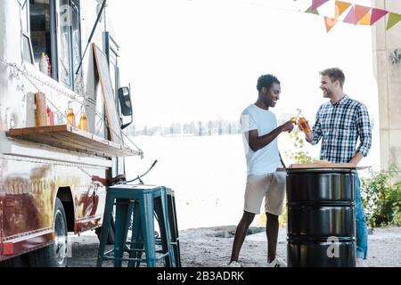des hommes multiculturels positifs clinquant des bouteilles de bière près d'un camion alimentaire à l'extérieur Banque D'Images