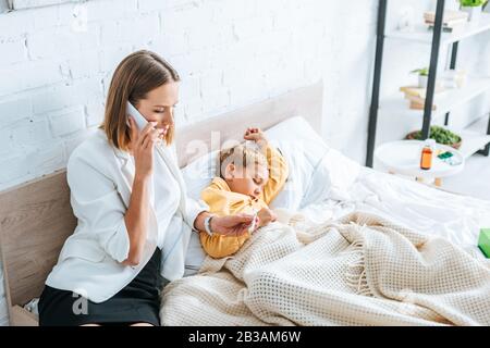 une femme inquiète à regarder un thermomètre et à parler sur son smartphone tout en étant assise près d'un fils malade Banque D'Images