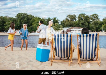 vue arrière de l'homme et de la femme assis dans des chaises longues près d'amis multiculturels s'amuser sur la plage Banque D'Images