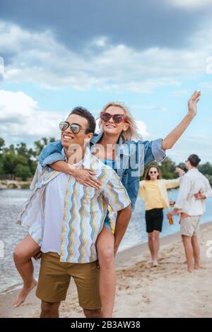 un homme de course souriant et souriant, qui soutient une petite amie tout en marchant sur la plage près d'amis multiculturels Banque D'Images