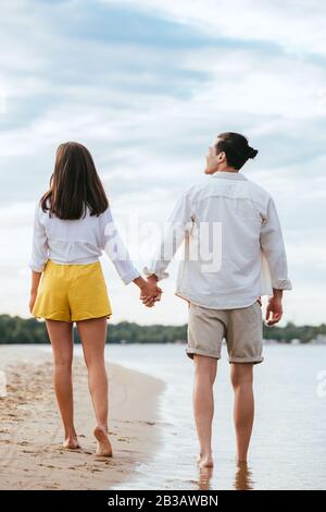 vue arrière du jeune couple tenant les mains et regardant l'un l'autre tout en marchant sur le bord de la rivière Banque D'Images