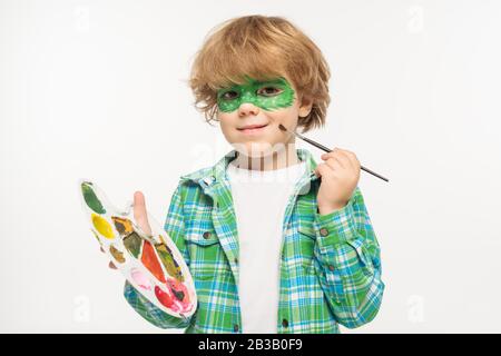 garçon gai avec masque de gecko peint sur le visage touchant la joue avec pinceau isolé sur blanc Banque D'Images