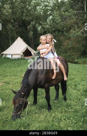 Cheval, deux filles enfants, sœurs, équitation un cheval à l'extérieur, souriant et s'embrassant. Filles équitation dans pré en été. Wigwam Banque D'Images