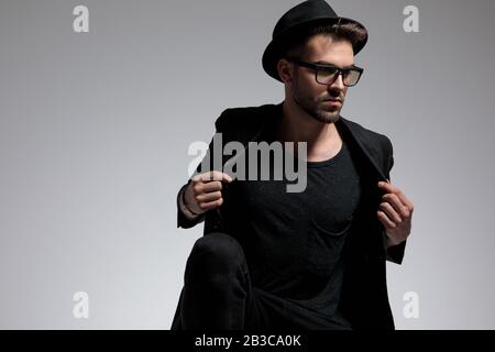 bon homme décontracté portant un chapeau et des lunettes cousue debout et veste de fixation sérieuse contre le fond gris de studio Banque D'Images