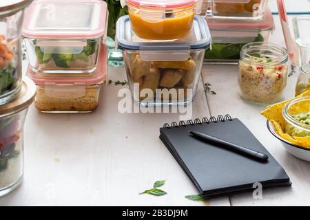 Scène de préparation de repas avec des tas de bocaux en verre remplis de repas maison. Un bloc-notes noir avec un stylo dessus est à l'avant. Banque D'Images