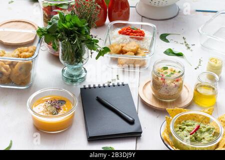 Scène de préparation de repas avec des tas de bocaux en verre remplis de repas maison. Un bloc-notes noir avec un stylo dessus est à l'avant. Banque D'Images