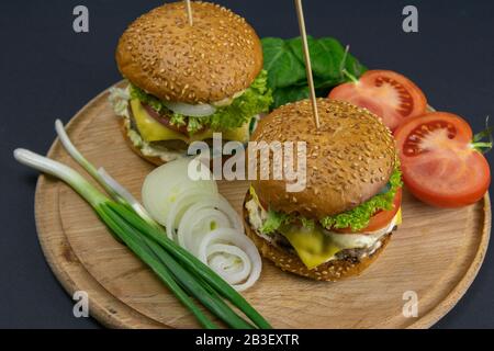 Deux hamburgers rapprochés sur une planche ronde en bois, à côté des tomates, des oignons hachés et des feuilles d'épinards Banque D'Images