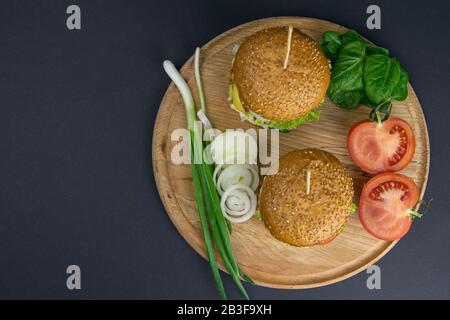 Deux hamburgers rapprochés sur une planche ronde en bois, à côté des tomates, des oignons hachés et des feuilles d'épinards Banque D'Images