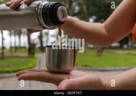 Une femme verse une boisson d'un thermos le tenant dans ses mains tout en se tenant dans un parc Banque D'Images