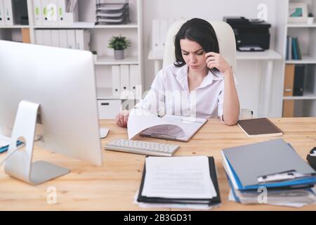 Jeune femme concentrée dans un chemisier blanc assis à un bureau en bois et des documents d'analyse dans le presse-papiers Banque D'Images