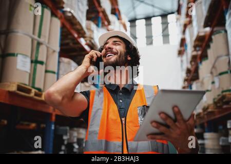 Vue à faible angle du superviseur d'entrepôt, à l'aide d'un téléphone, pour la communication, tenant une tablette numérique avec casque et gilet de sécurité Banque D'Images