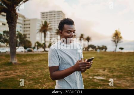 Portrait d'un jeune athlète souriant se tenant dans le parc le matin textant des messages sur le téléphone portable hi Banque D'Images